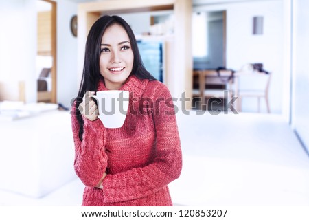 Beautiful happy smiling woman dressed with winter clothes holding a hot cup of coffee at home