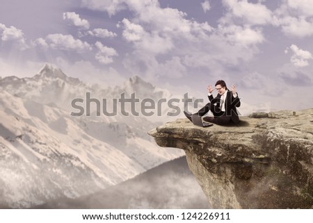 Businessman is sitting happily while working on his laptop on top of a mountain