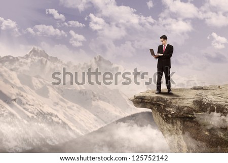 Businessman holding a laptop on top of a mountain under cloudy sky