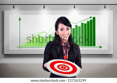 Happy businesswoman holding dartboard while standing in front of profitable bar chart