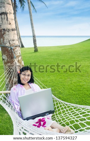 Asian student with laptop on hammock at beach, Indonesia