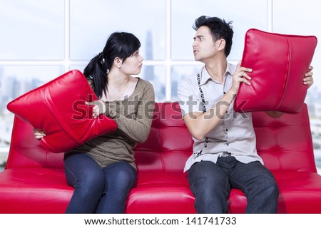 Angry couple on red sofa fighting with pillow at apartment