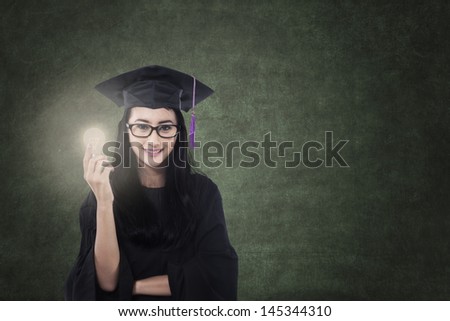 Attractive Asian graduate holding lit bulb with blank blackboard in class