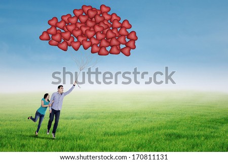 Happy couple holding heart shaped balloons and flying over a green meadow