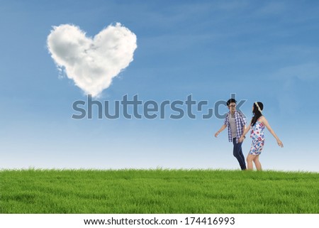 Romantic young couple walking together on the meadow while holding hands with heart shaped cloud