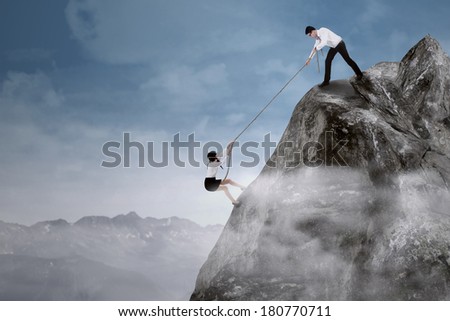 Portrait of businessman help his partner to climb the cliff by pulling her with a rope