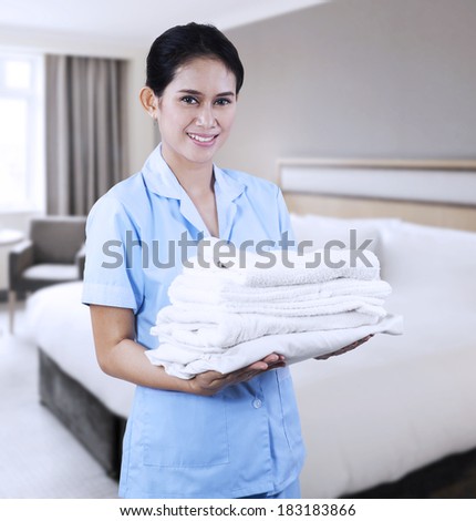 Smiling young cleaning lady holding towels shooting at hotel room