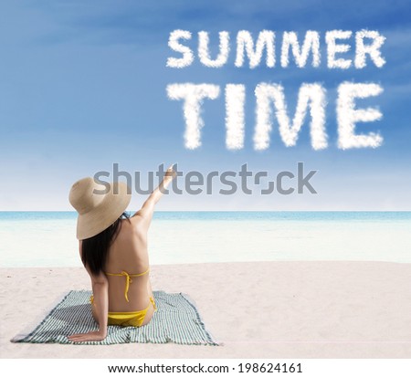 Portrait of smiling beautiful woman wearing bikini sitting on mat and pointing at a text of summer time