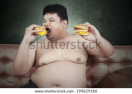 Greedy fat man eating hamburger while watching tv at home