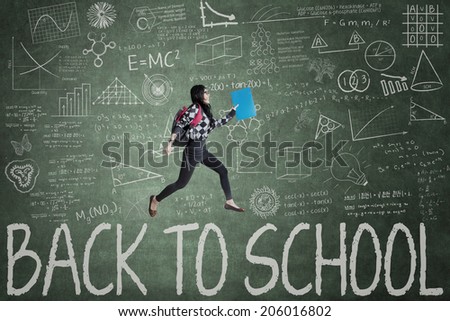 Female student jumping in classroom through back to school text on blackboard