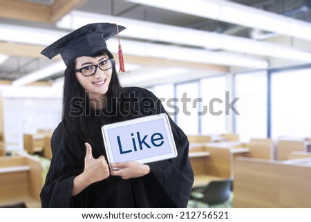 Young woman with graduation gown in library showing a like text on digital tablet