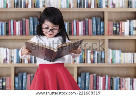Asian little girl reads book seriously in library with bookcase background
