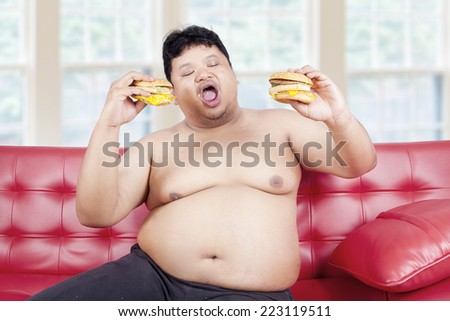 Portrait of overweight man sitting on sofa and eat two burgers at home