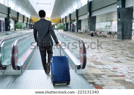 Rear view of businessman carrying luggage walk toward escalator in airport hall