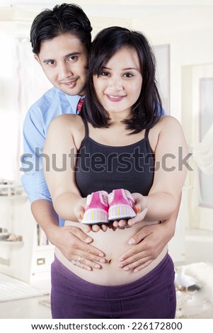 Portrait of happy parents standing in bedroom and showing pair of pink shoes for baby girl
