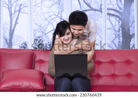 Portrait of young man standing on the back of sofa and looking his wife using laptop while hugging her