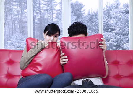Portrait of scared couple watching horror movie at home and hiding on the pillows