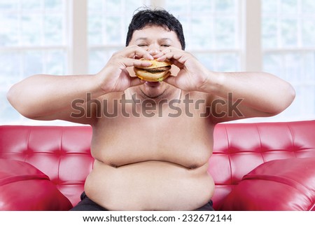 Fat man eating hamburger seated on sofa at home