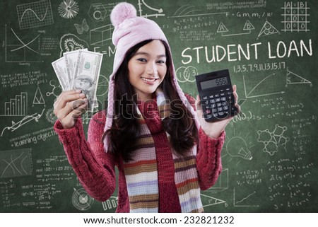 Portrait of beautiful female student wearing winter clothes and holding money with calculator in class