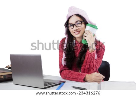 Teenage girl wearing a winter knitted clothes, doing schoolwork and smiling at the camera while holding a cup of warm coffee