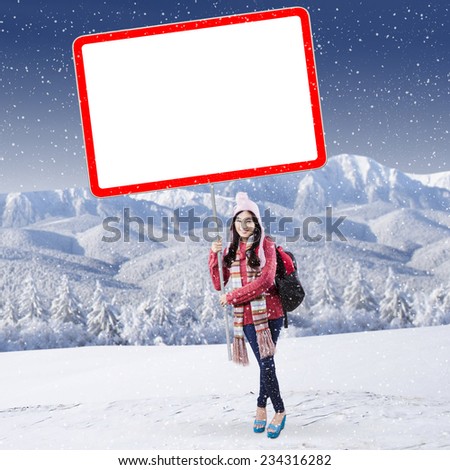 Cheerful female student in winter clothes holding blank board at snowy mountain