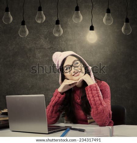 Female student studying with laptop and textbooks, wearing winter clothes and looking at bright lamp