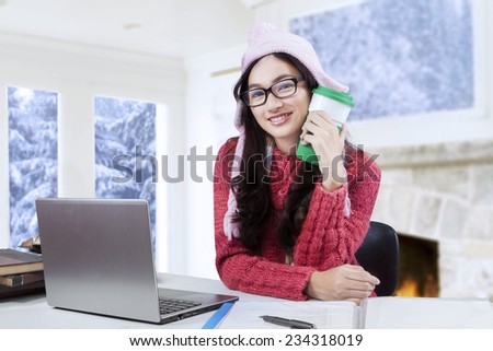 Young girl smiling at the camera while holding a cup of warm drink and studying on the table at home