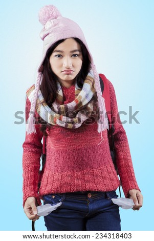 Worried schoolgirl in winter clothes showing empty pockets over blue background