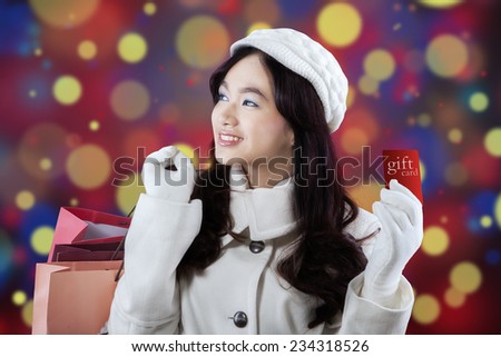 Lovely girl smiling happy while holding a gift card and shopping bags over light christmas background