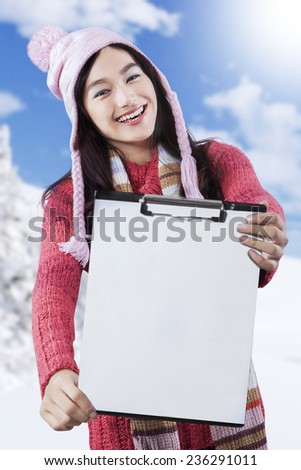 Young asian girl wearing a knitted winter clothes, showing blank clipboard at the camera