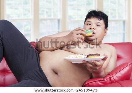 Portrait of lazy overweight person eating donuts while sitting on the sofa at home