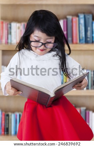 Cute little girl standing in the library while reading a textbook