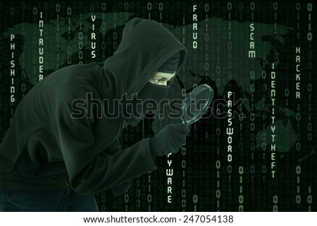Male hacker wearing black mask and uses magnifying glass for looking binary code