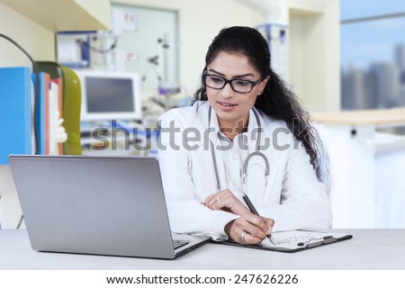 Portrait of young indian doctor working in the hospital while looking at laptop and write on clipboard