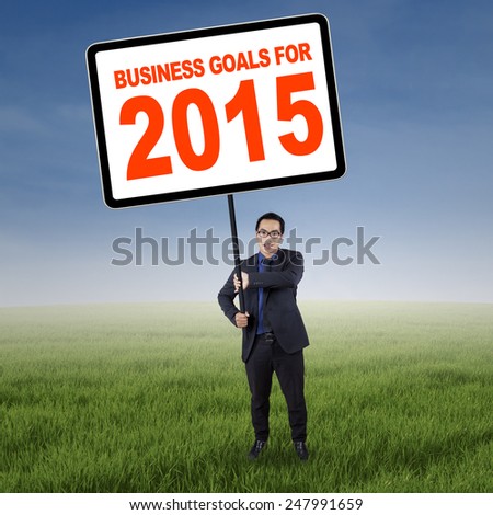 Young entrepreneur standing at field and holding a board with a text of business goals for 2015