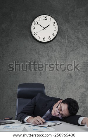 Young caucasian businessman sleeping on desk with clock on the wall
