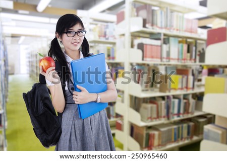 Young attractive female student standing in the library while holding a red apple and folder