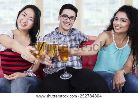 Three mixed race people sitting on sofa while toast with champagne and smiling at the camera