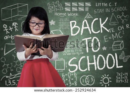 Portrait of little student standing in front of blackboard while reading a book with a text of back to school
