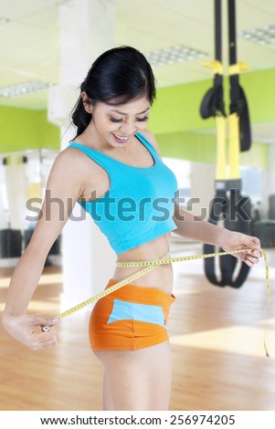 Young asian woman standing in the fitness center while measuring her waistline with measuring tape
