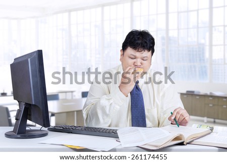 Male entrepreneur sitting in the office while looking at the book and biting a burger