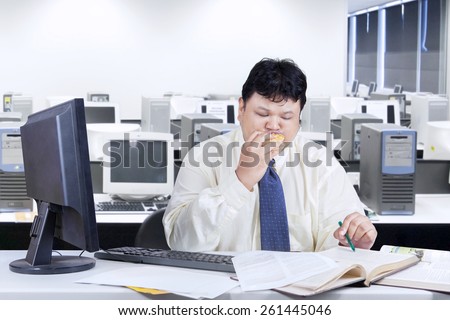 Obese entrepreneur working on table while biting a burger, shot in the office