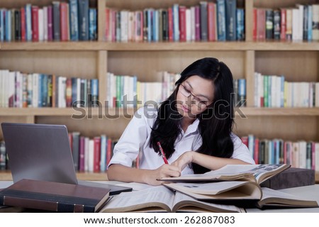 Teenage girl studying with textbooks while writing on a book in the library