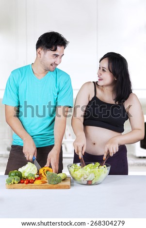 Portrait of cheerful pregnant woman and her husband preparing salad in the kitchen