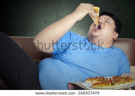 Obese person eats pizza while sitting on couch at home