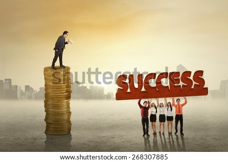 Young business leader standing on the stack of golden coins while giving order to his employees