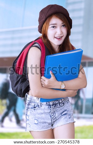 Teen girl with casual clothes and holds a folder standing at schoolyard while smiling on the camera