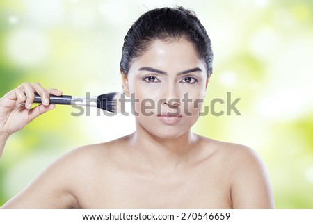 Attractive young female model with black hair and healthy skin, using makeup brush on her face