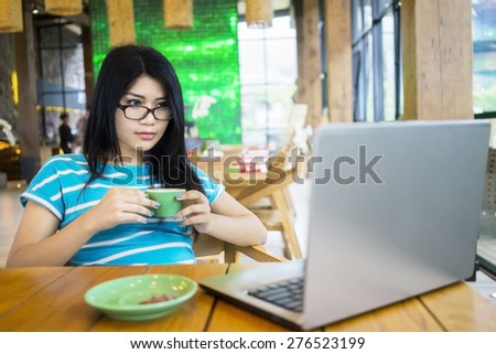 Portrait of young woman sitting in the cafe while holding a cup of coffee with laptop on the table