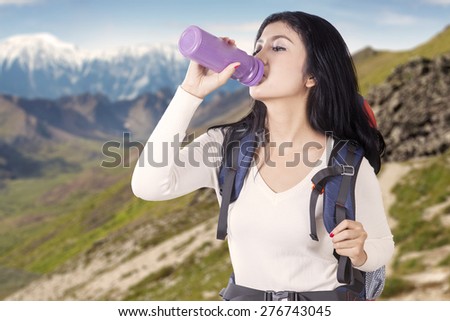 Portrait of female backpacker drinks fresh water from the bottle while carrying backpack on the edge of mountain